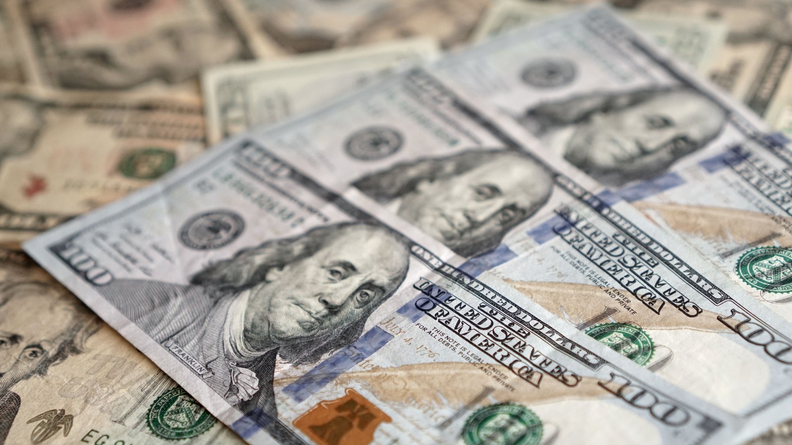 United States one hundred dollar bills lying overlapped and face up, filling the whole frame, with the portrait of Benjamin Franklin sharp in the centre and the words UNITED STATES OF AMERICA legible across two of the notes. No other lettering, no logo and no person appear in the picture.