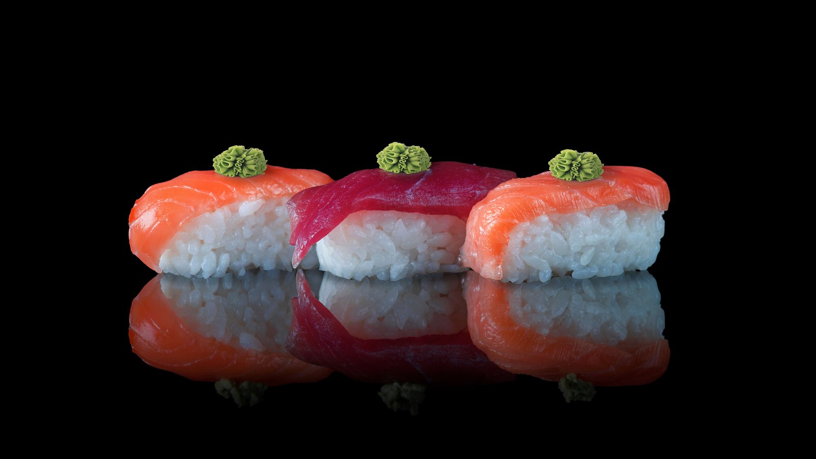 Three pieces of nigiri sushi standing in a row, two topped with salmon and the middle one with tuna, each finished with a small mound of grated wasabi, mirrored in the black reflective surface they stand on against a black background.