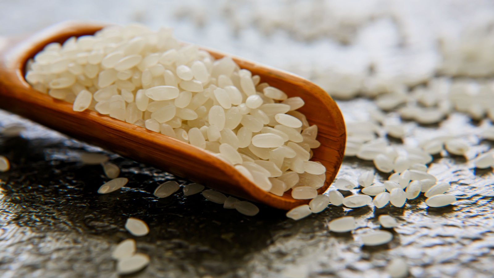 A close macro of short-grain white rice heaped on a wooden scoop, with loose grains scattered across the dark textured surface beneath it.