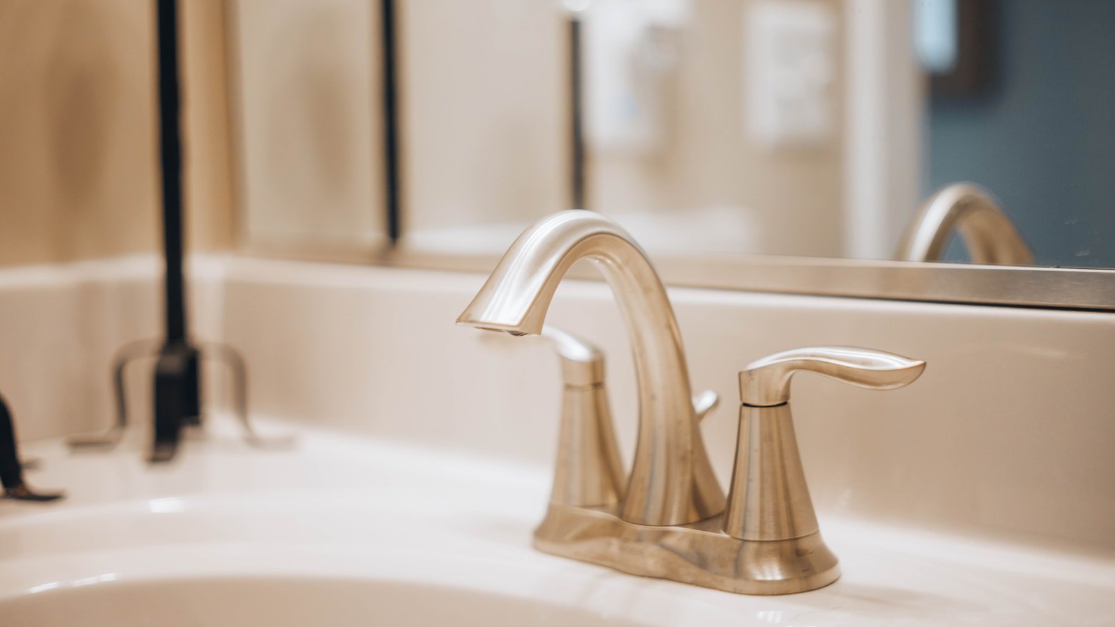 A brushed metal bathroom faucet above a white sink, with a mirror reflecting the fixture behind it.