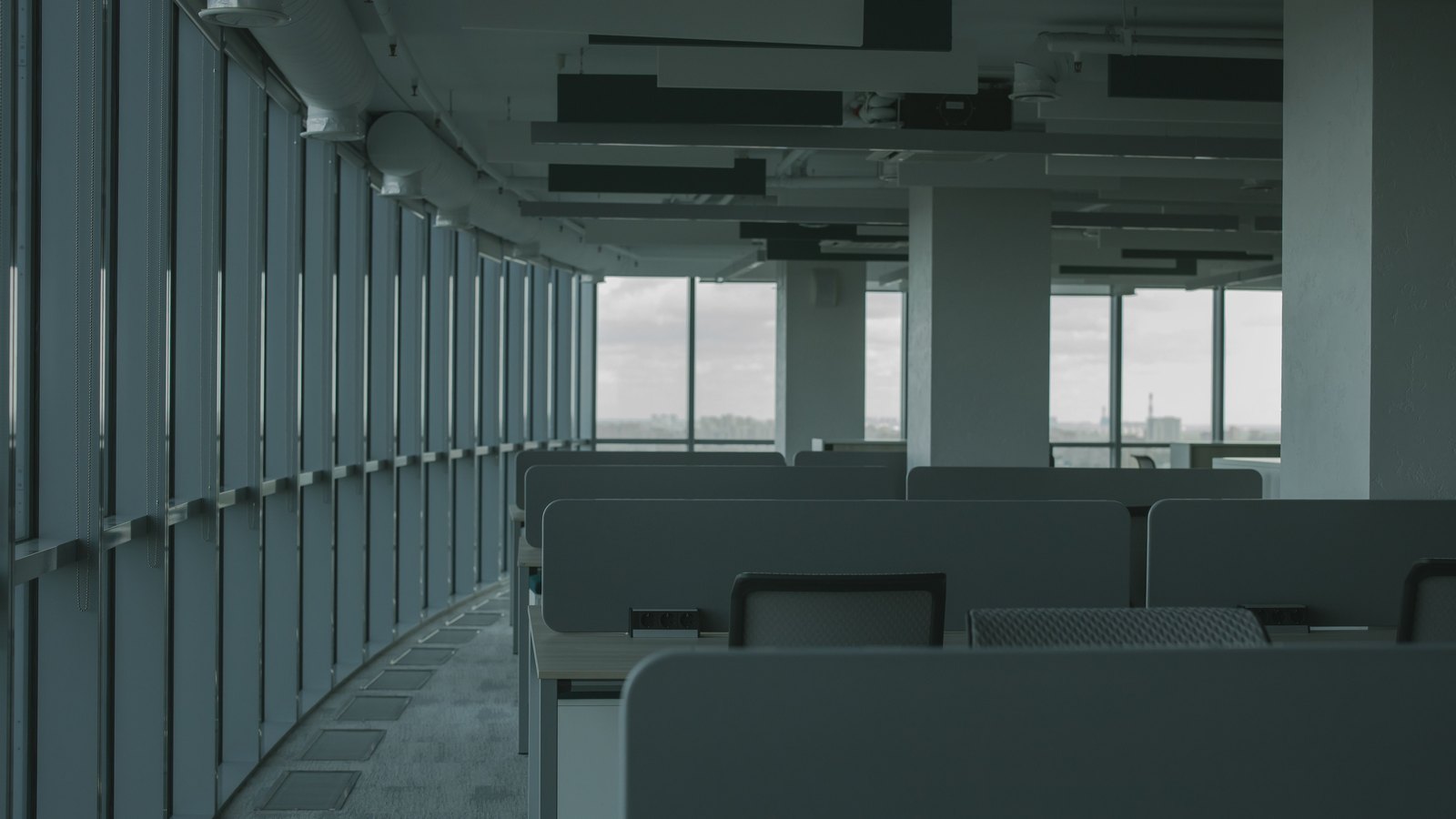 Rows of unoccupied workstations in an office with large windows.