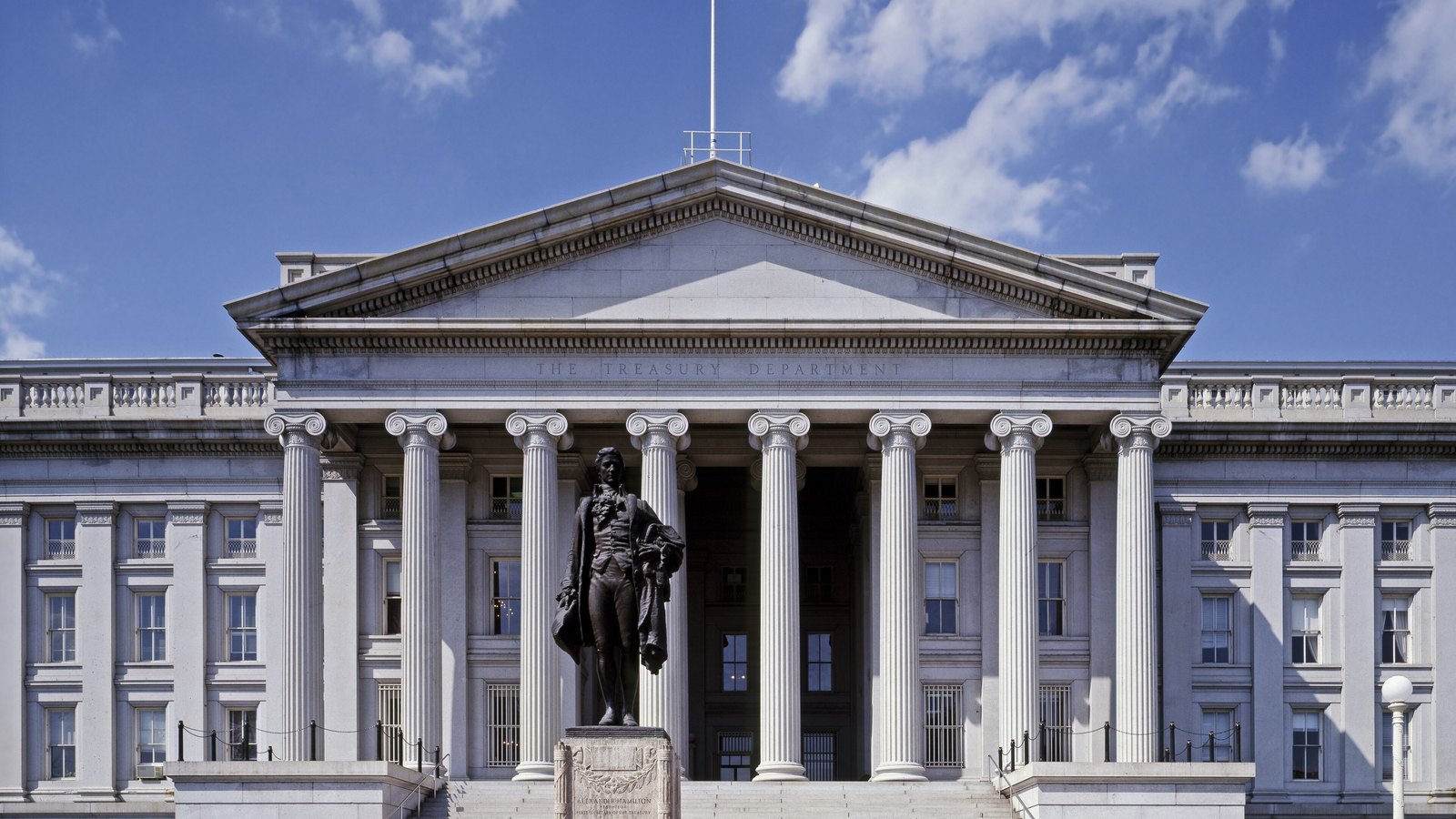 The colonnaded north front of the United States Treasury Building in Washington under a blue sky, the entablature carved with the words The Treasury Department, and the bronze statue of Alexander Hamilton standing on its plinth in front of the central portico.