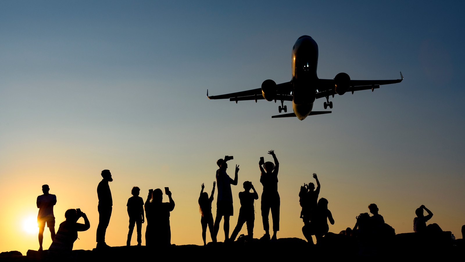 A passenger aircraft passes above silhouetted onlookers at sunset