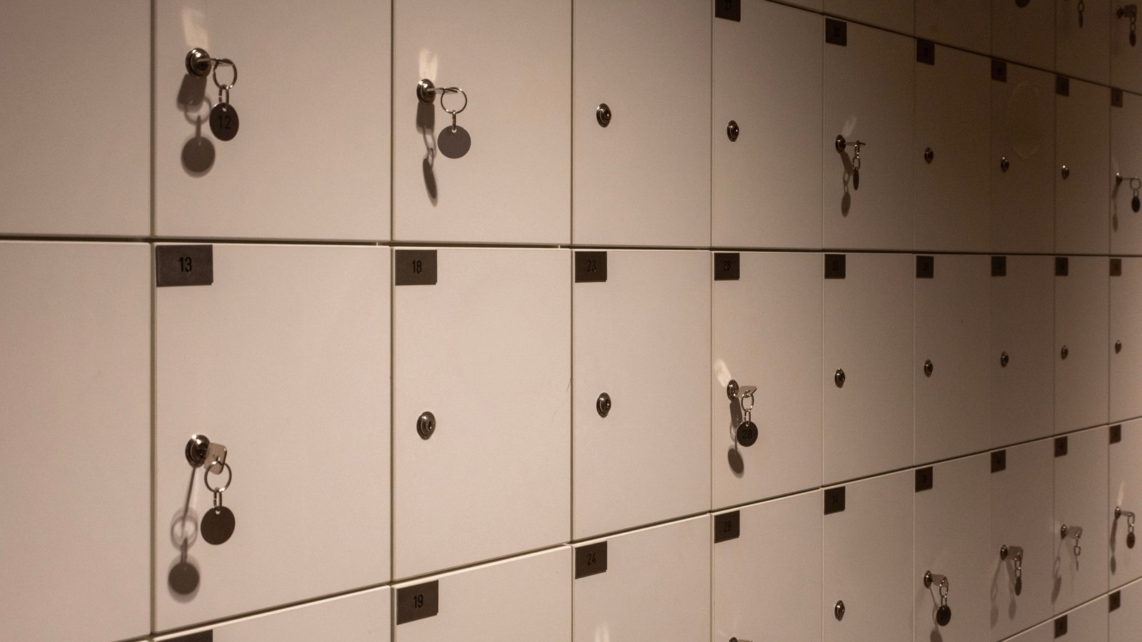 A wall of small square white locker doors receding to the right, each with a round lock and a numbered plate, several with a key and a plain metal tag still hanging in the lock, lit warmly from one side. There is no person, no lettering beyond the door numbers, no logo, no brand and no place cue anywhere in the frame.