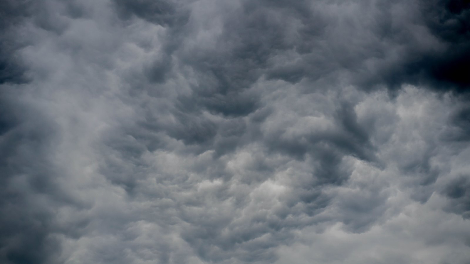 A dense grey overcast filling the whole frame, layered turbulent cloud in charcoal and pale grey with ragged lobes hanging from the underside and a darker bank across the upper right. The frame holds cloud only. No horizon, land, water, people, buildings, signage or text are in view.