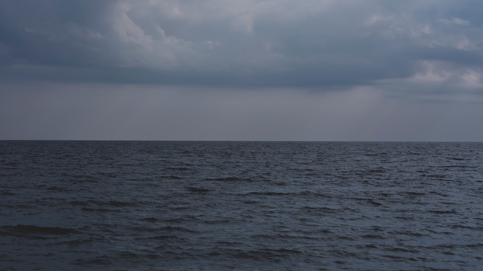 A dark blue open ocean running flat to the horizon under a heavy grey deck of storm cloud, with rain hanging below the cloud base. There is no land, no vessel, no person, no signage and no legible text anywhere in the frame, and nothing in it identifies a place. A generic illustration of a storm over open water, not a photograph of this event.
