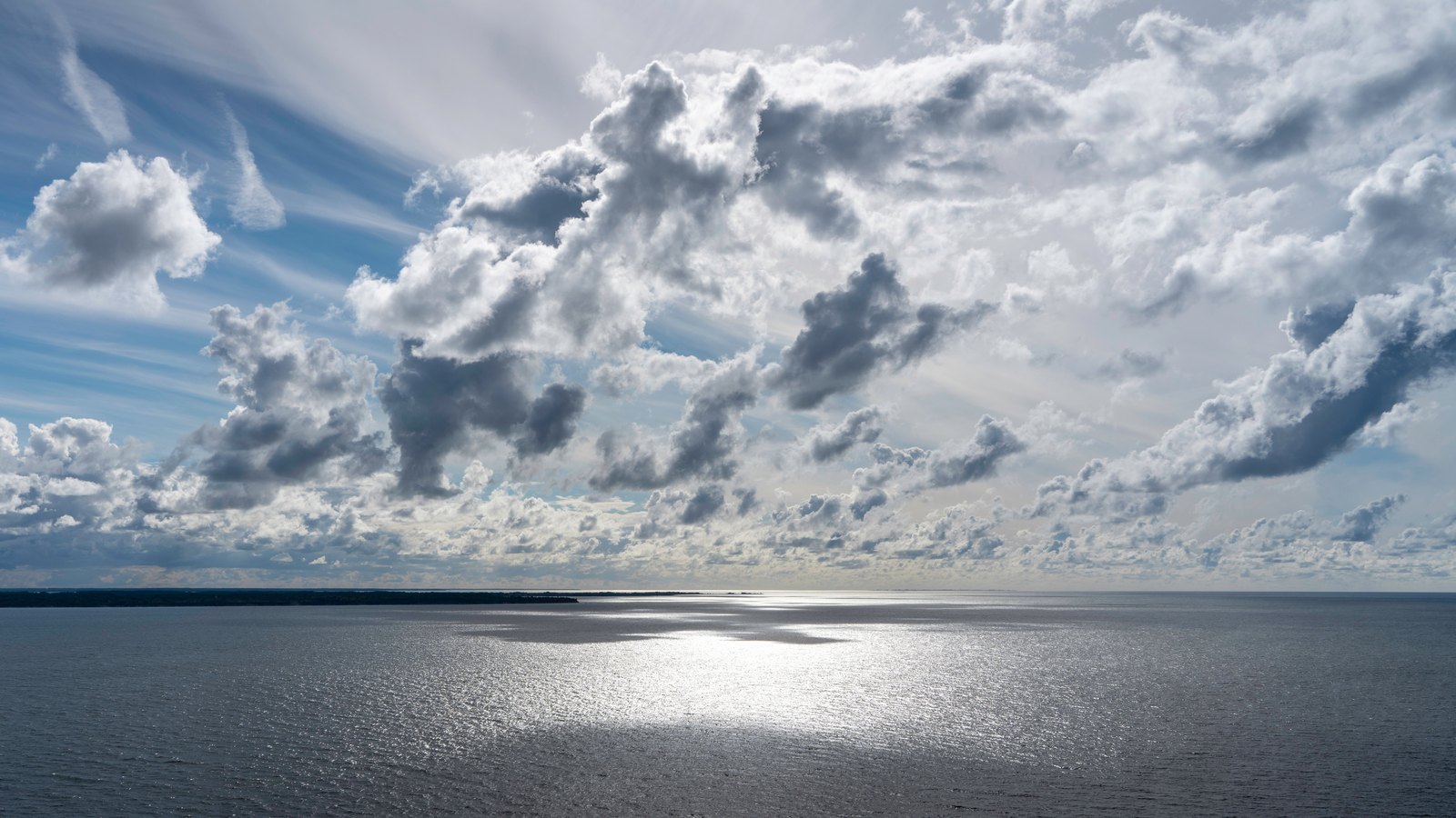 A wide stretch of calm grey water under a broken sky, with scattered cumulus cloud lit from behind and thin cirrus streaking the blue at upper left. Sunlight glints in a bright path across the middle of the water. A low dark strip of distant shoreline runs along the horizon at the far left. No people, boats, buildings, signage or text are in view.