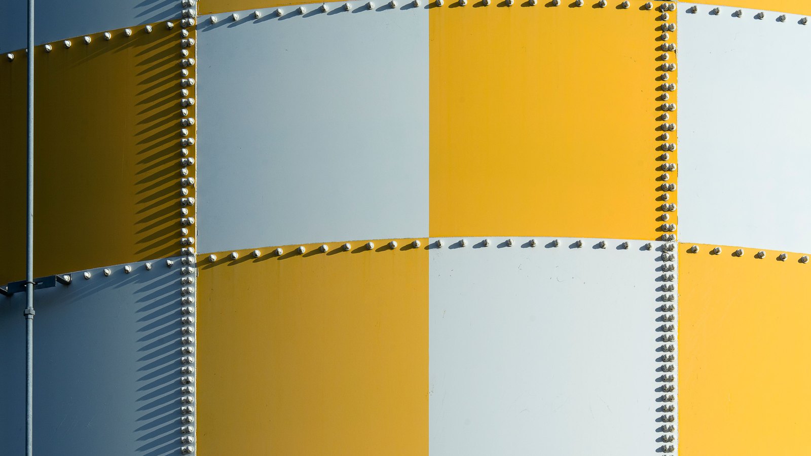 A close photograph of the curved wall of a riveted metal structure filling the whole frame, painted in a checkerboard of yellow and off white square panels, with rows of small domed rivet heads running along every seam and casting short shadows. A thin vertical pipe runs down the left edge. No sky, horizon, text, brand mark, person or identifiable place is in view.