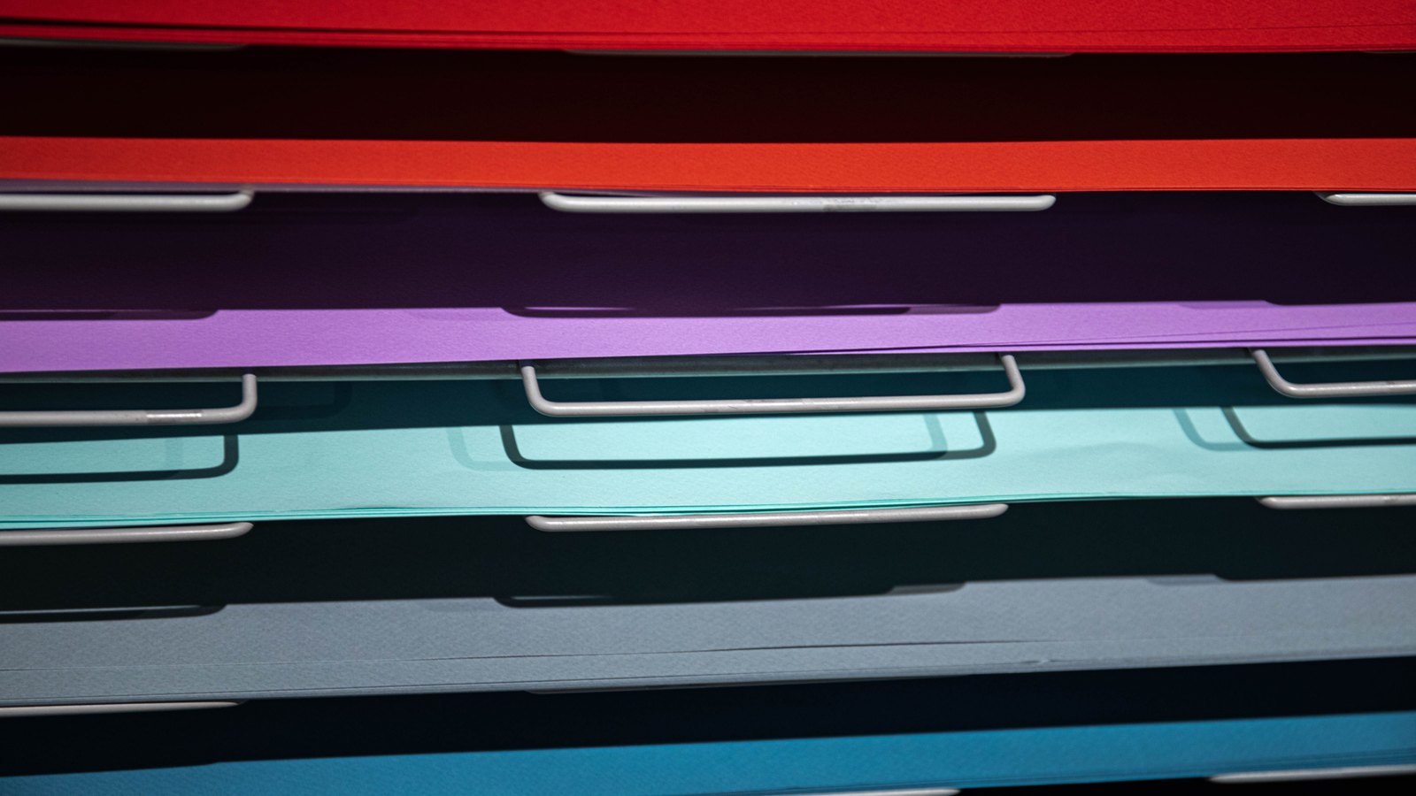 Sheets of coloured paper resting on the steel rails of a filing rack, seen edge on.
