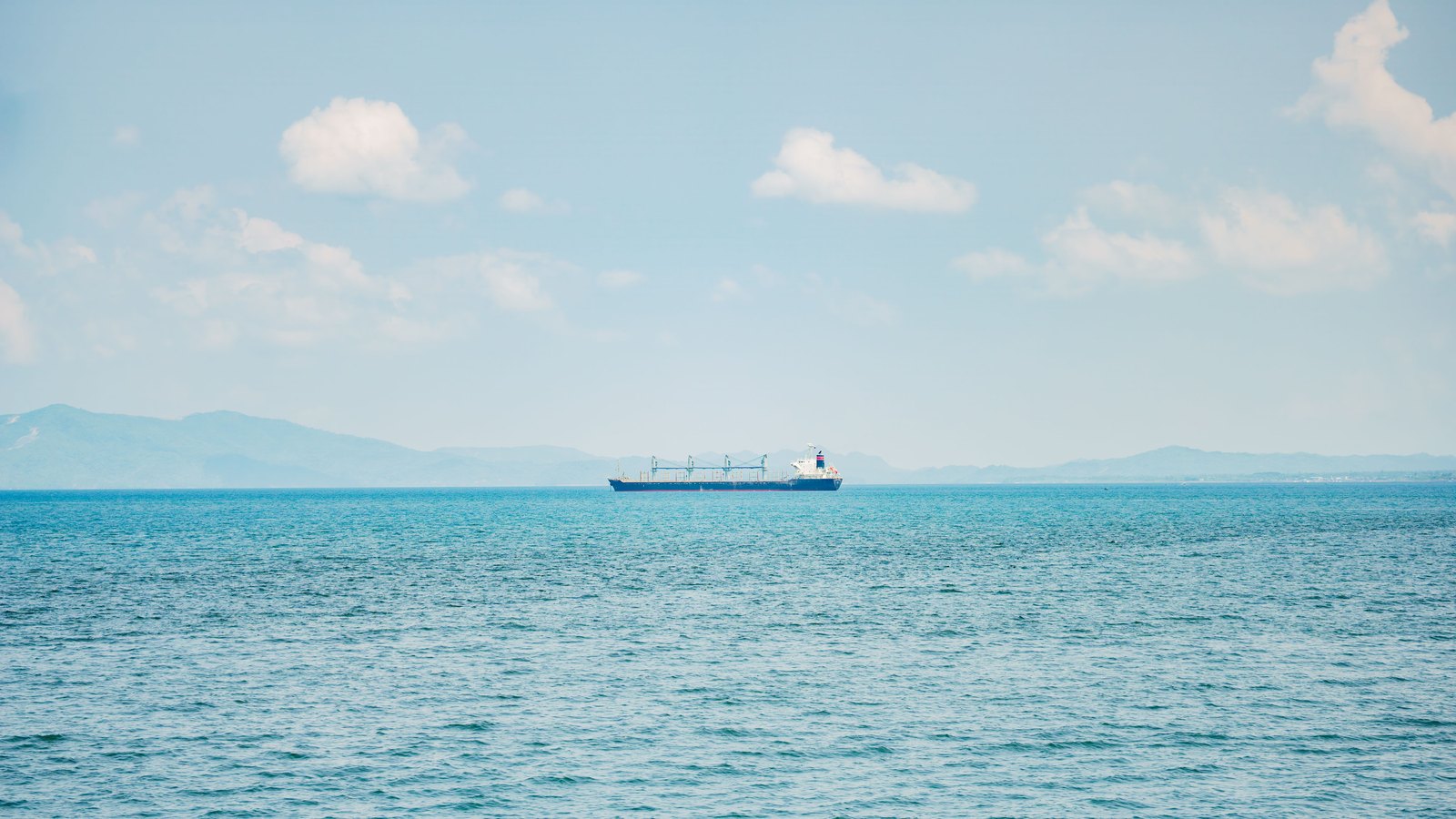 A distant cargo ship crosses calm blue water below a pale sky, with mountains on the horizon.