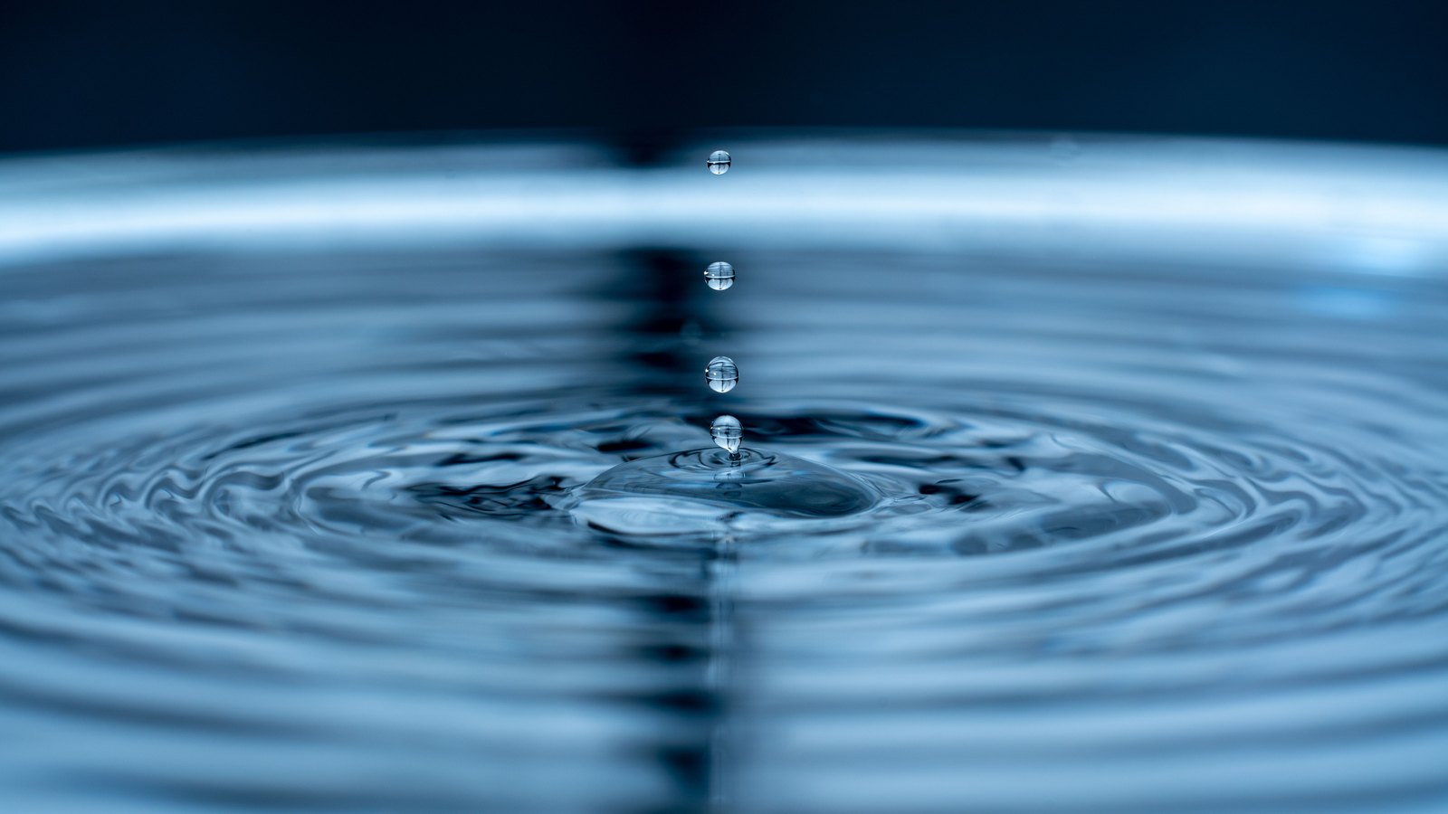 A close photograph of blue water filling the whole frame, lit from behind, with a raised dome at the centre where a drop has struck and concentric rings running out from it to every edge. Four small round droplets hang in a vertical line above the impact point. No text, brand mark, person, place or object edge is in view.