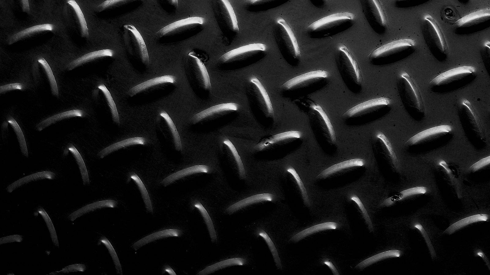 A close photograph of dark steel chequer plate lit hard from one side, rows of raised oval treads set on the diagonal catching a bright specular highlight against an almost black ground. No text, lettering, brand mark, product, tool, person or place is in view.