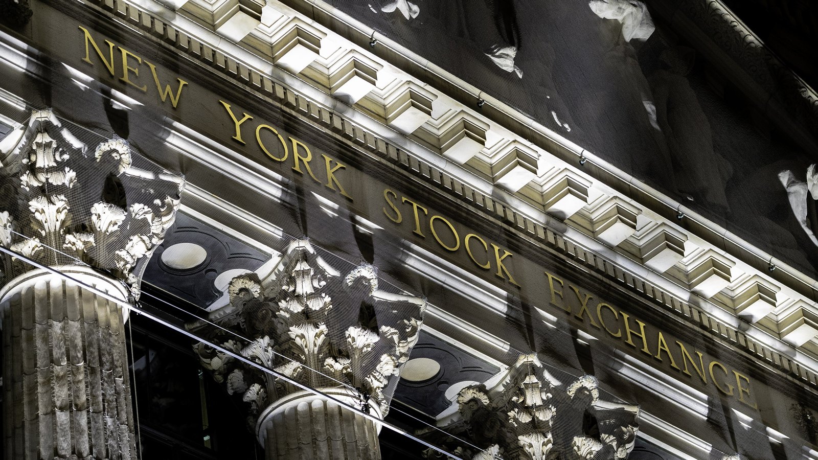A close view of the entablature of the New York Stock Exchange building on Broad Street, the words NEW YORK STOCK EXCHANGE in gold lettering above Corinthian capitals, seen at an angle through the protective mesh that covers the facade. No person is in the frame.