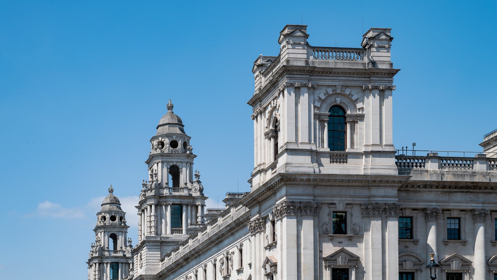 The upper storeys of the Portland stone facade of the Government Offices Great George Street in Whitehall, London, which houses HM Treasury, showing a square corner tower with an arched window and three domed stone cupolas receding along the roofline against a clear blue sky. No people, vehicles, signage or lettering are in the frame.