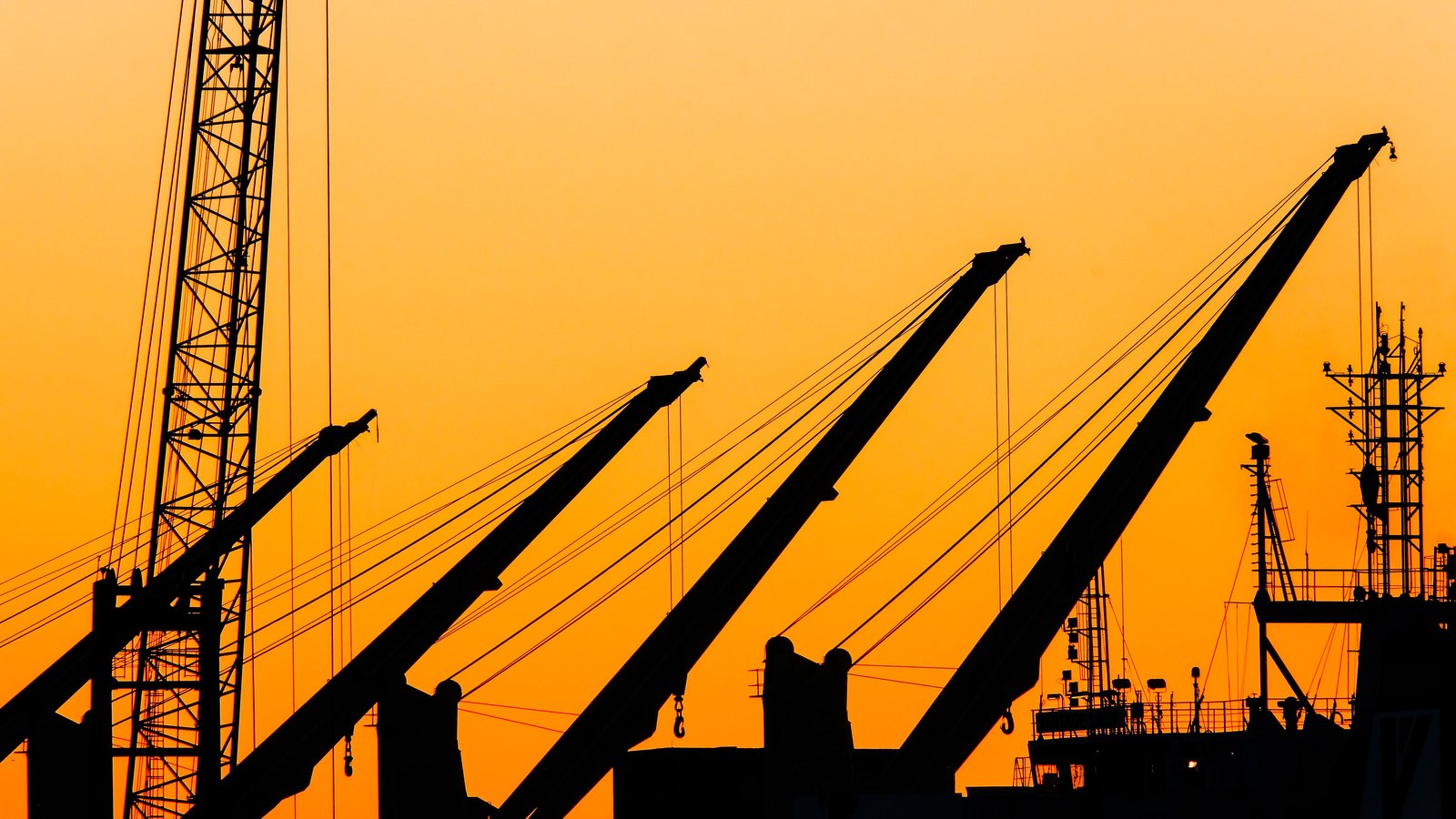 Cargo cranes and a ship stand in silhouette against an orange sunset sky.