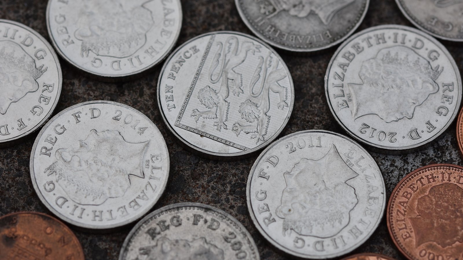 Worn British coins, ten pence and penny pieces bearing Queen Elizabeth II, scattered on a dark stone surface.
