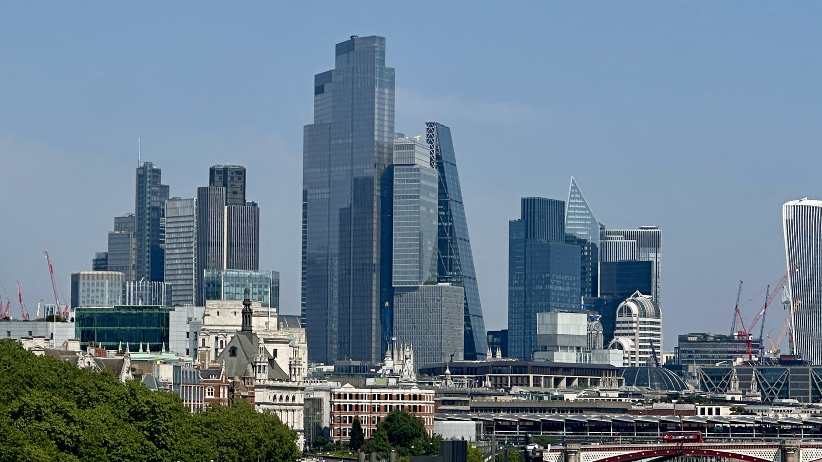 Glass office towers of the City of London under a clear blue sky, with older stone and brick buildings, construction cranes and trees in the foreground and a red arched bridge along the bottom edge. No legible text or identifiable person is in view.