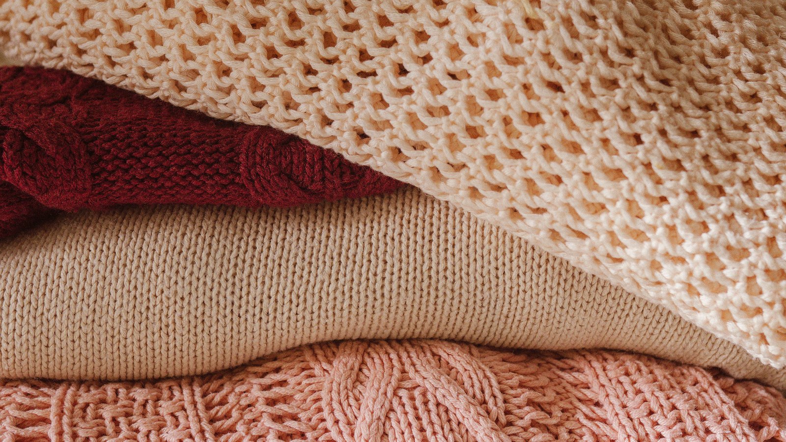 A close view of a stack of folded knitted sweaters in cream, deep red, beige and pink, their cable and open stitches filling the frame. No labels, text or people are in view.