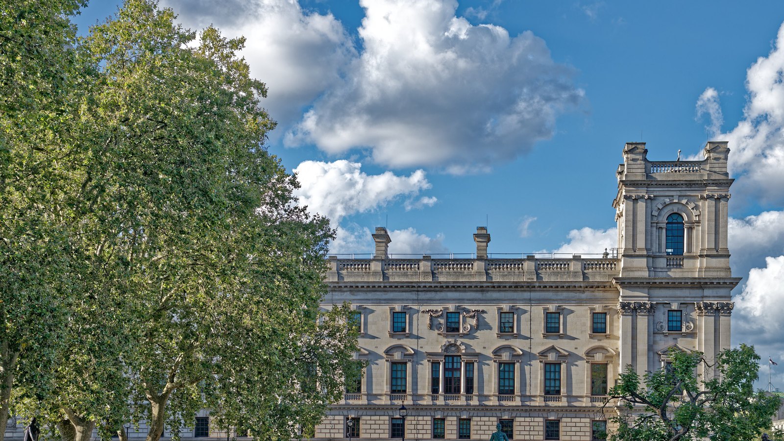 The upper storeys and towers of the HM Treasury building in London rise behind trees beneath a blue sky.