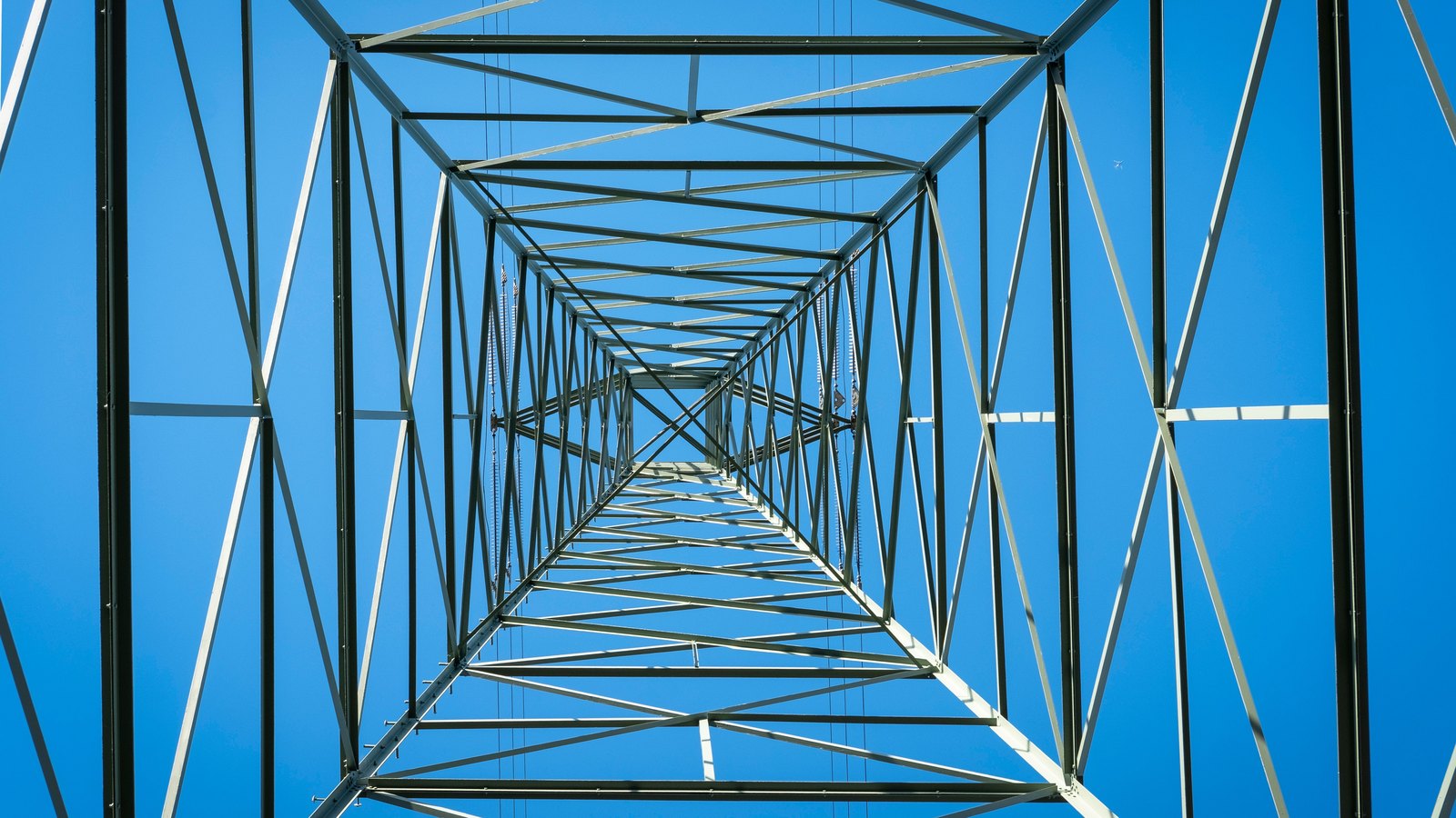 Steel transmission pylon photographed straight up from its base, lattice bracing converging against a clear blue sky.