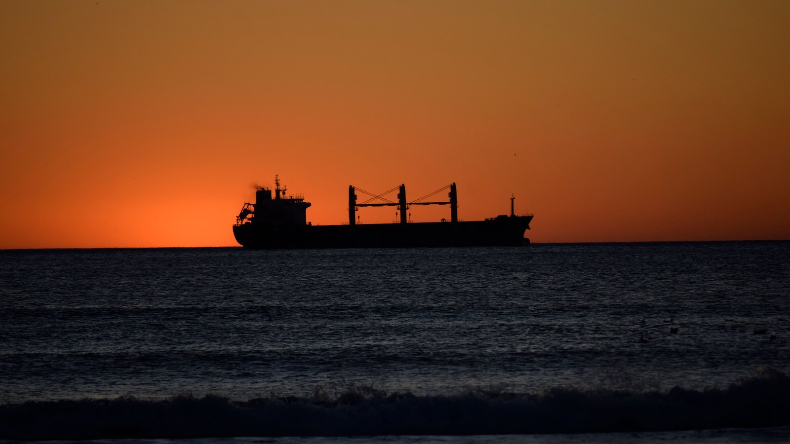 A cargo ship appears in silhouette on the horizon against an orange sunset over dark water.