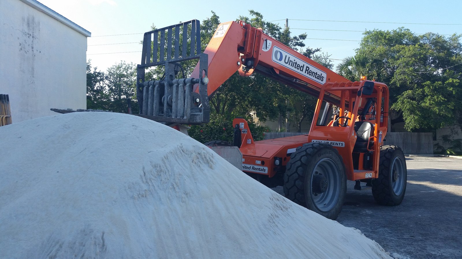 An orange United Rentals telehandler with its fork carriage raised stands on a paved lot behind a large mound of pale sand, with trees and a white wall behind it.