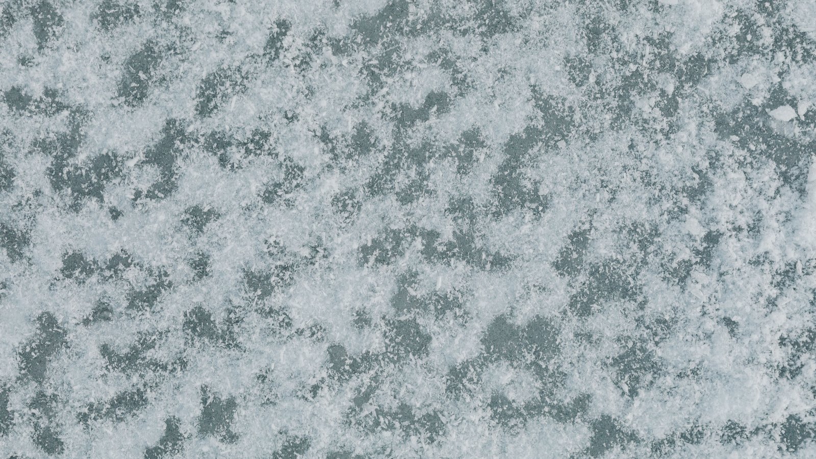 Fresh snow crystals scattered across a dark grey-green frozen surface and filling the whole frame. No horizon, footprint, tree, object or lettering is in the picture.
