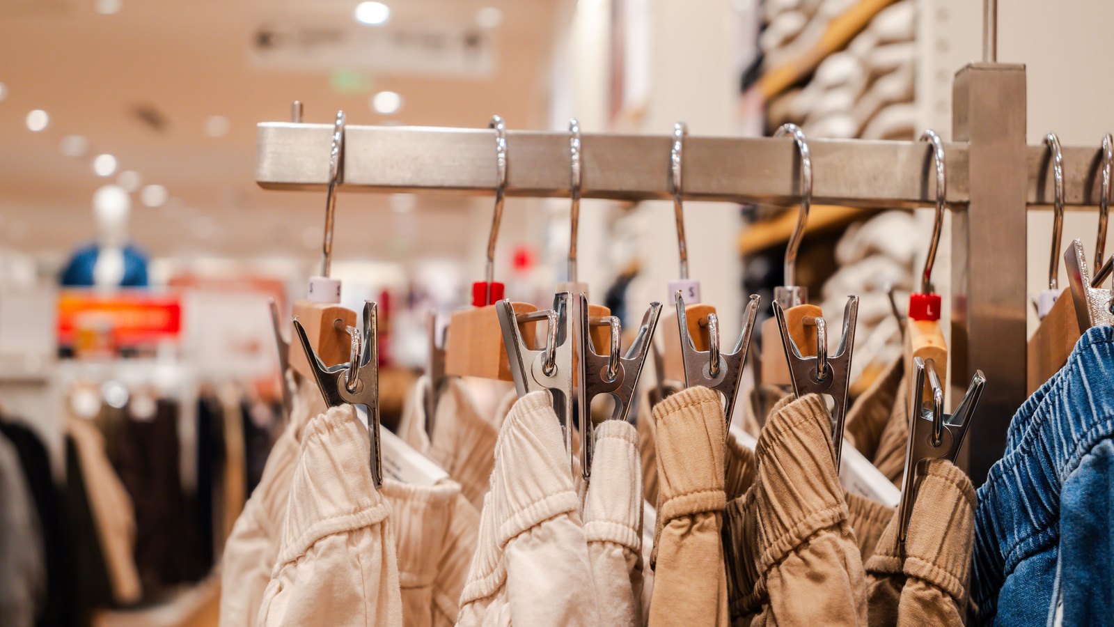 A steel rail in a clothing shop hung with clip hangers, each gripping the waistband of a pair of trousers in cream, oatmeal and tan cotton, with one pair of blue denim at the right edge. Small white size tabs read S and L. The rest of the shop behind is thrown completely out of focus.