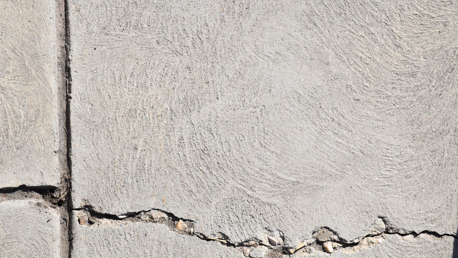 A close crop of a pale grey concrete surface in daylight, its face carrying the fine swirled marks of a broom finish. A straight joint runs down the left of the frame and a ragged crack runs across the lower third, with small pieces of aggregate and grit exposed along its edge. No people, vehicles, buildings, signage, lettering or brand marks are visible, and no place can be identified from it. Pexels titles it a cracked concrete wall. An abstract surface used as a plain illustration, not a photograph of US-41, of North Fort Myers or of this rollover, and not evidence about the condition of any road.