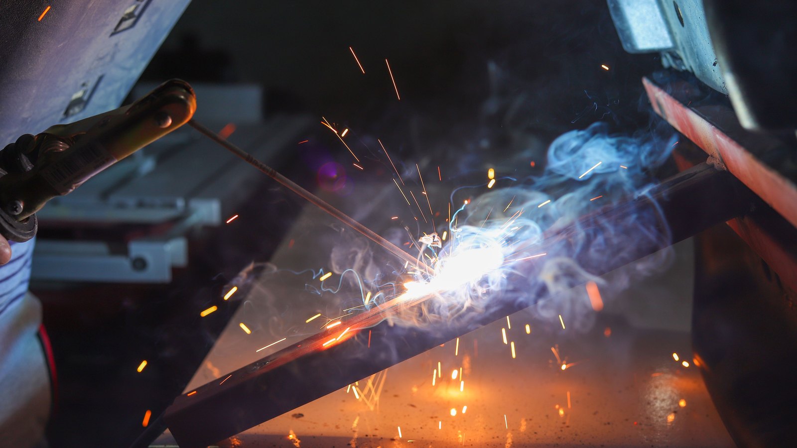 Bright welding sparks and smoke surround a metal workpiece in a factory.