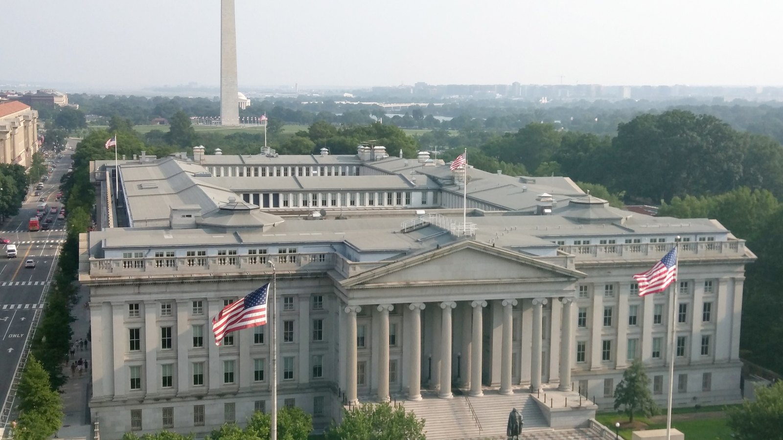 The pale stone United States Treasury Building seen from above, with American flags along its columned front and the Washington Monument behind it.