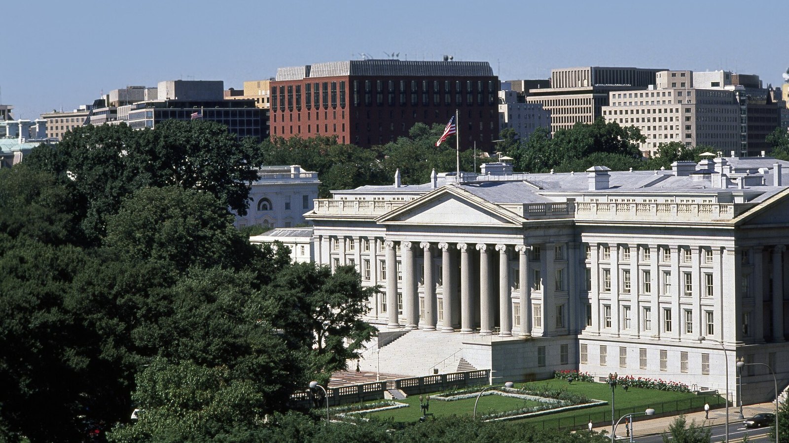 The United States Treasury Building in Washington, seen from above with its south portico, pale stone walls and an American flag.