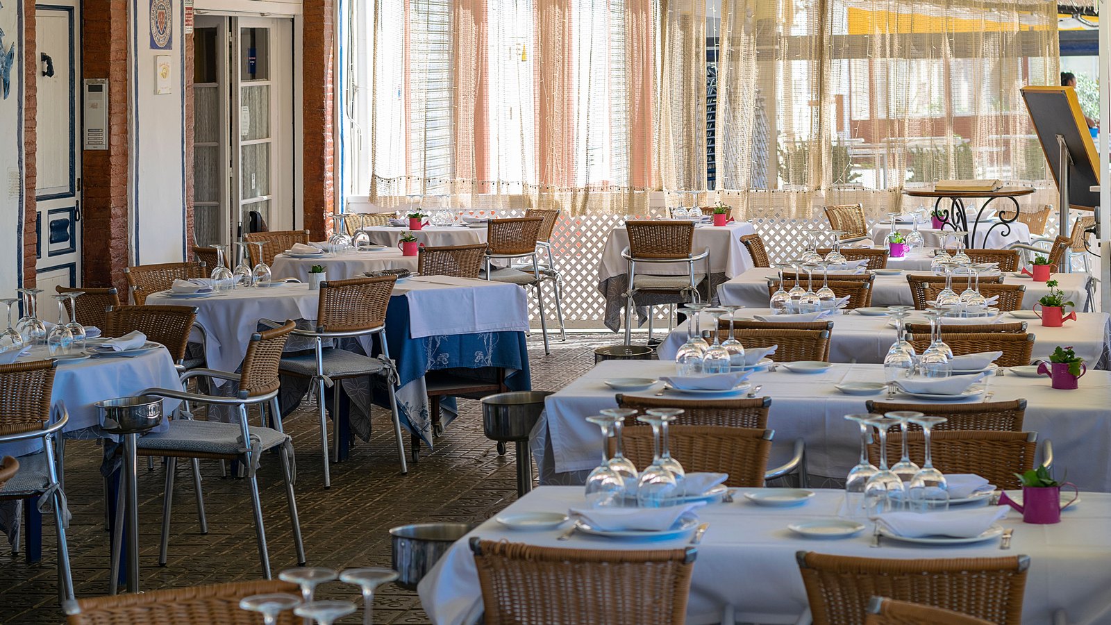 An empty restaurant dining room set with white tablecloths, wicker chairs, glassware and small potted plants in daylight.