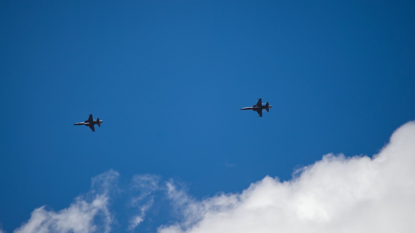 Two fighter jets cross a blue sky above white clouds, not a photograph of this event.
