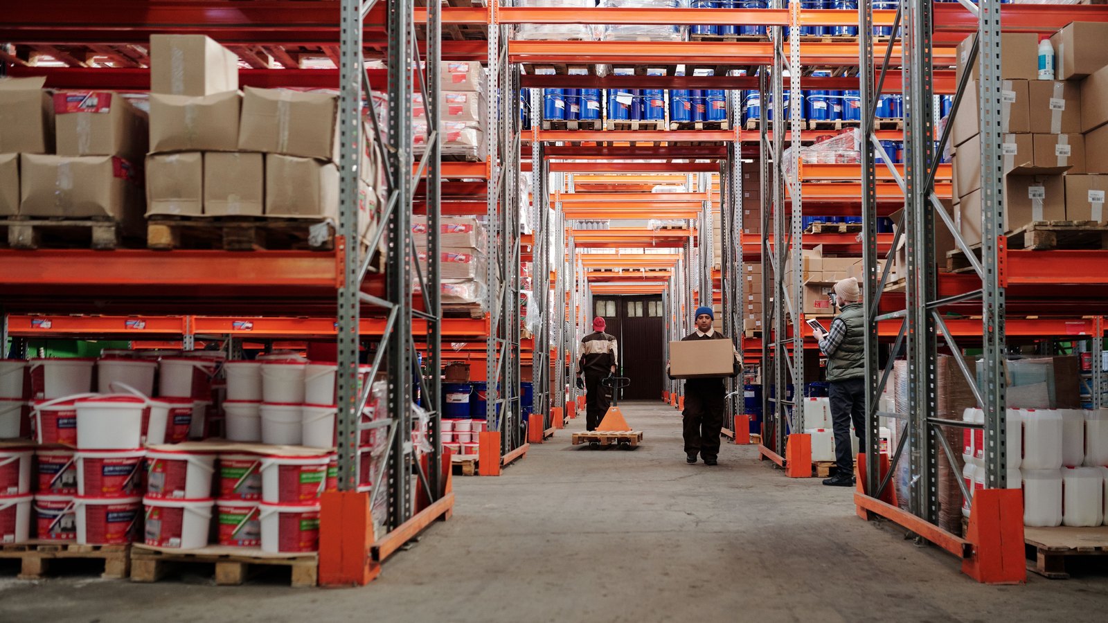 Workers move boxes through a warehouse lined with stocked pallet racks.