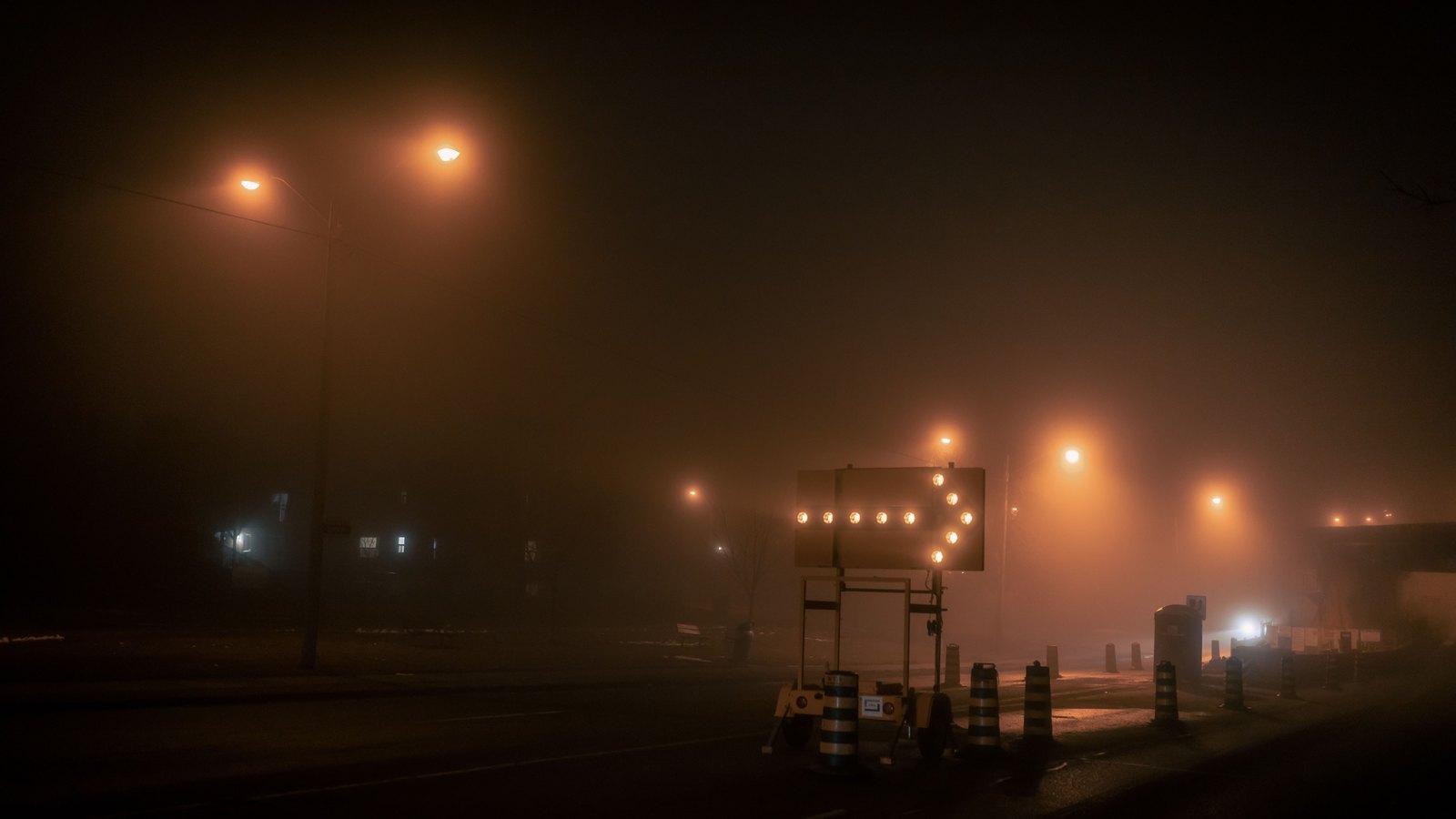 An illuminated lane direction arrow and traffic barrels on a foggy road at night, not a photograph of this event.
