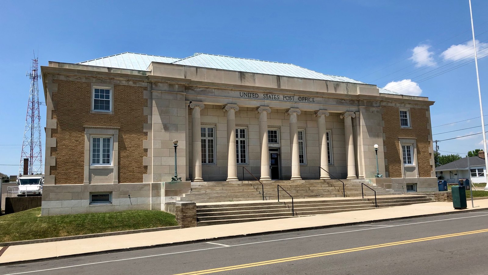 The United States Post Office in Greensburg, Indiana, a brick and limestone building fronted by six Ionic columns, with the words UNITED STATES POST OFFICE cut into the stone above them and two blue collection boxes standing on the pavement at the right.