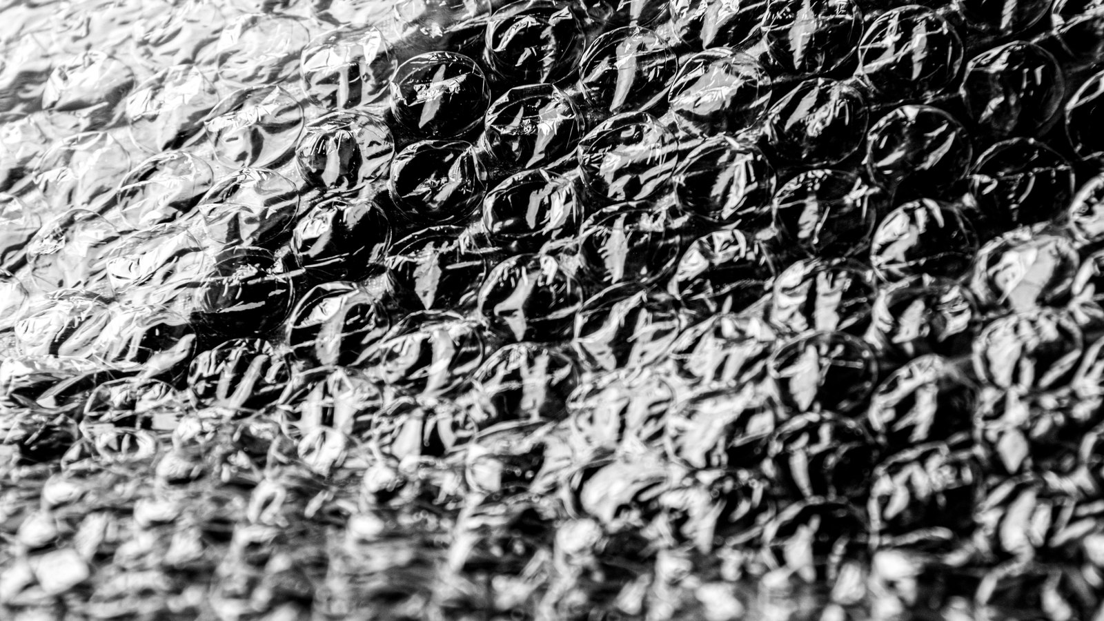 A sheet of bubble wrap photographed in black and white, rows of glossy air pockets filling the frame, the plastic creased and catching hard highlights across the domes.