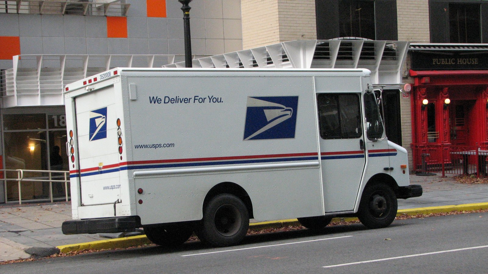 A white United States Postal Service delivery van parked at a city kerb and seen from the rear quarter, carrying the blue and white sonic eagle logo on the side and on the rear door, the line We Deliver For You, the address www.usps.com and a red and blue stripe along the body. Fallen leaves lie in the gutter and a red painted shopfront stands on the pavement behind.