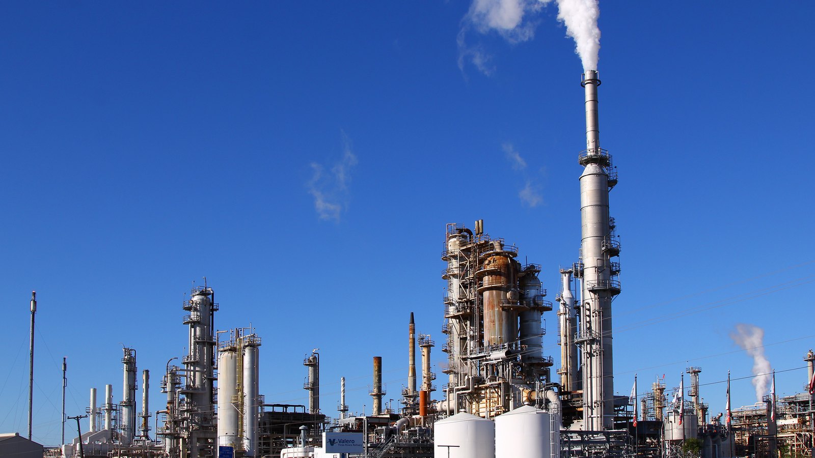 Distillation columns, storage tanks and a tall stack releasing white vapour at Valero's refinery in Three Rivers, Texas, with a Valero sign at the gate under a deep blue sky.