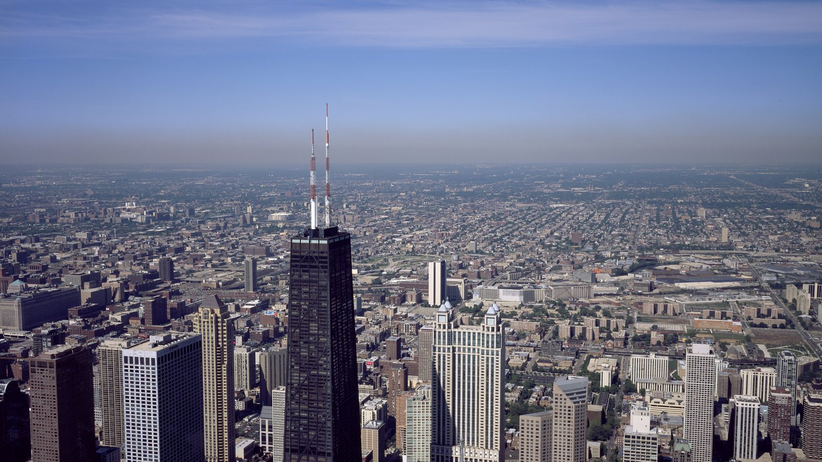An aerial view over Chicago on a clear day, the John Hancock Center and its twin antennas in the foreground and the low rise city stretching to the horizon behind it.
