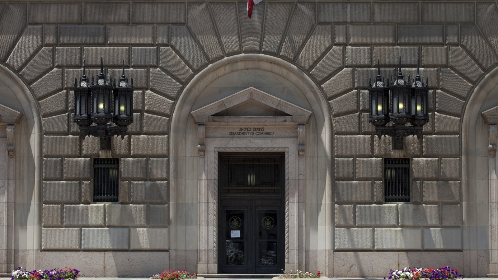 The arched limestone entrance of the Herbert C. Hoover Building in Washington, with UNITED STATES DEPARTMENT OF COMMERCE carved on the lintel above dark metal double doors that carry the department's gold seal, a large wrought iron lantern on a bracket to either side, and beds of red and purple flowers along the pavement below.