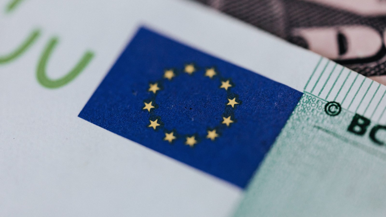 A close macro of a euro banknote, showing the blue rectangle with the twelve gold stars of the European flag, the green edge of a large numeral behind it and part of the printed lettering at the right.