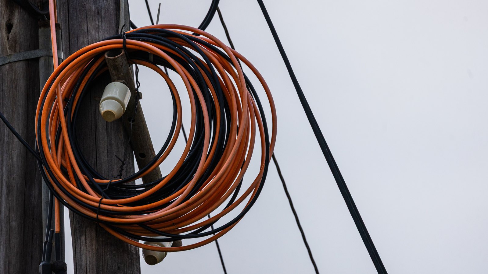 A coil of surplus orange and black cable strapped to a weathered wooden utility pole, two white ceramic insulators behind it, and taut spans of black line running away across a flat white overcast sky.