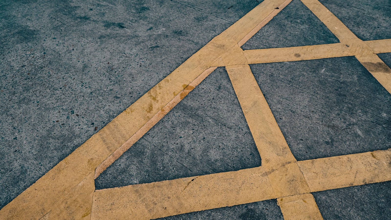 A close crop of a grey concrete surface photographed at an angle, crossed by broad bands of worn yellow paint meeting at angles to form a hatched pattern, the paint chipped and stained and the concrete flecked with dark aggregate. No text, signage, brand mark, vehicle, person or identifiable place is visible, and no horizon. A plain marked surface used as an illustration, not a photograph of Veterans Parkway, of the vehicle, or of this crash or its scene.
