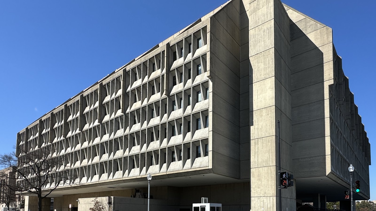 A long concrete government building in Washington photographed from a street corner against a clear blue sky. Its facade is a repeating grid of angled precast fins set over recessed windows, and the whole slab is lifted above an open colonnade at ground level. Bare winter branches stand at the left and a lamp post and pedestrian signal at the right. No names or lettering are legible.