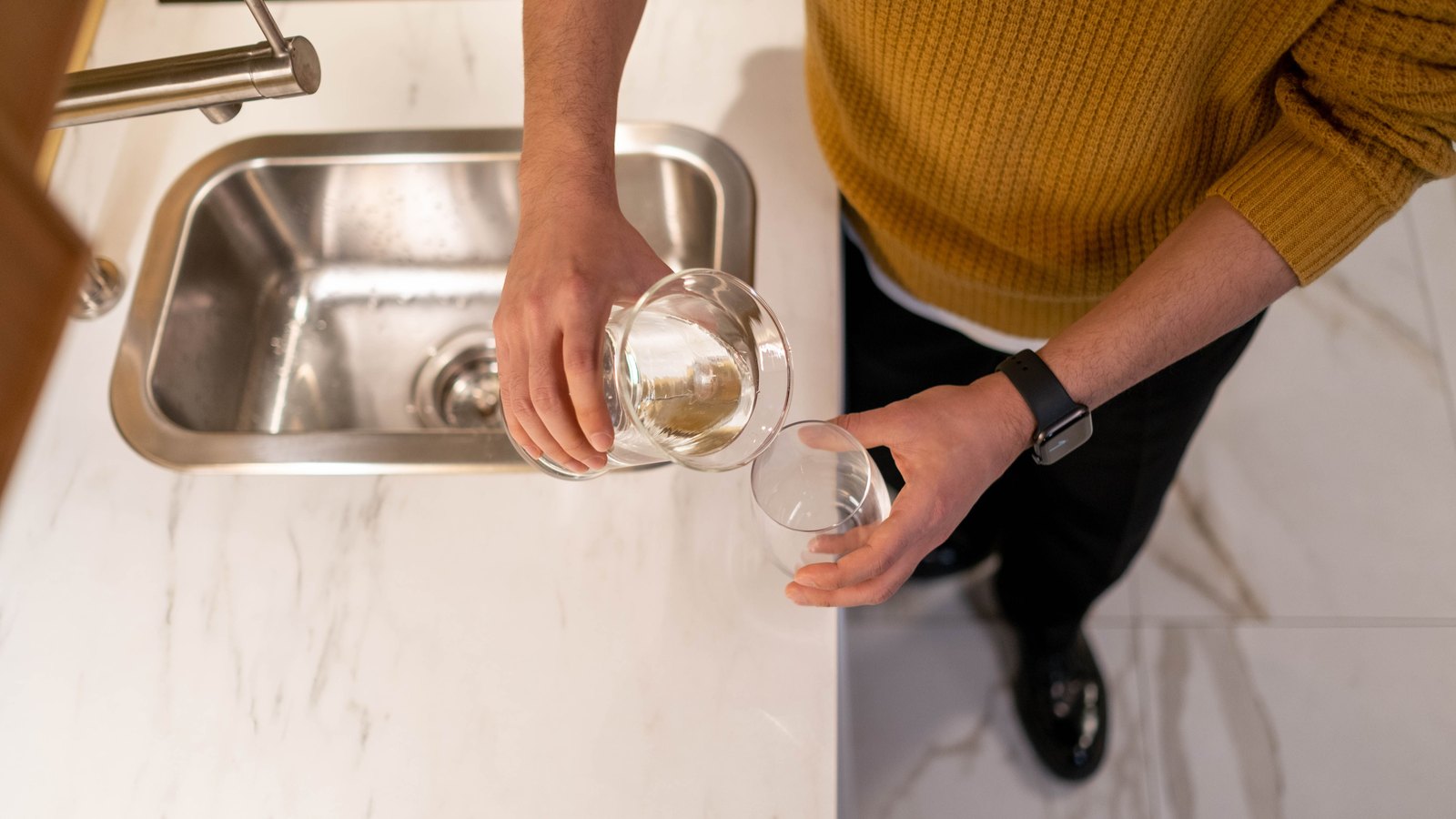Seen from above, a person in a mustard yellow sweater stands at a white marble kitchen counter beside a small stainless steel sink, pouring clear water from a glass carafe held in one hand into a short drinking glass held in the other. A brushed steel mixer tap sits at the edge of the sink. No face is visible and no text, signage or branding appears anywhere in the frame. Stock photograph, not a photograph of this event.