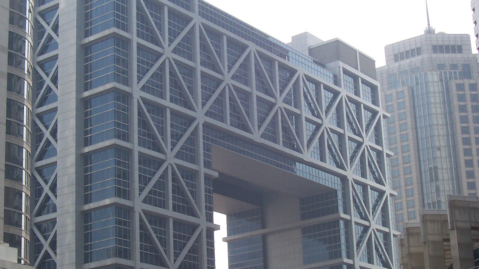 The Shanghai Stock Exchange building in Lujiazui, a grey concrete frame of large diagonal braces enclosing a blue glass tower, with a suspended block bridging the open gap at its centre and other Pudong towers standing behind it under a white sky.