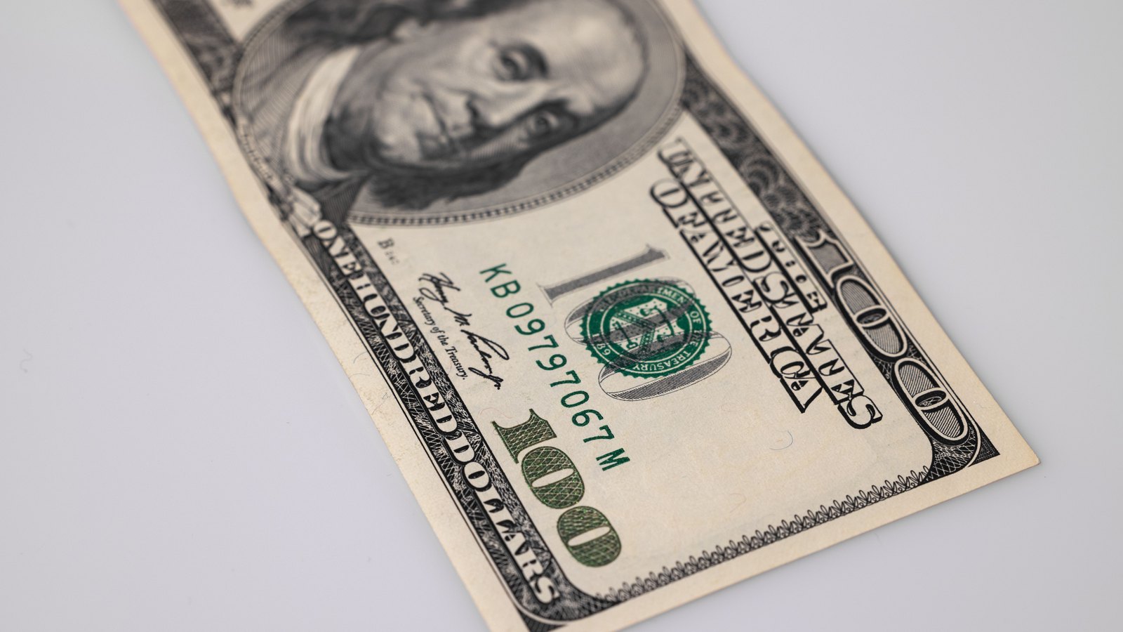 A single United States one hundred dollar note of the older design lying face up but inverted on a plain white surface, the portrait of Benjamin Franklin at the top of the frame and the green Treasury seal and serial number across the middle.