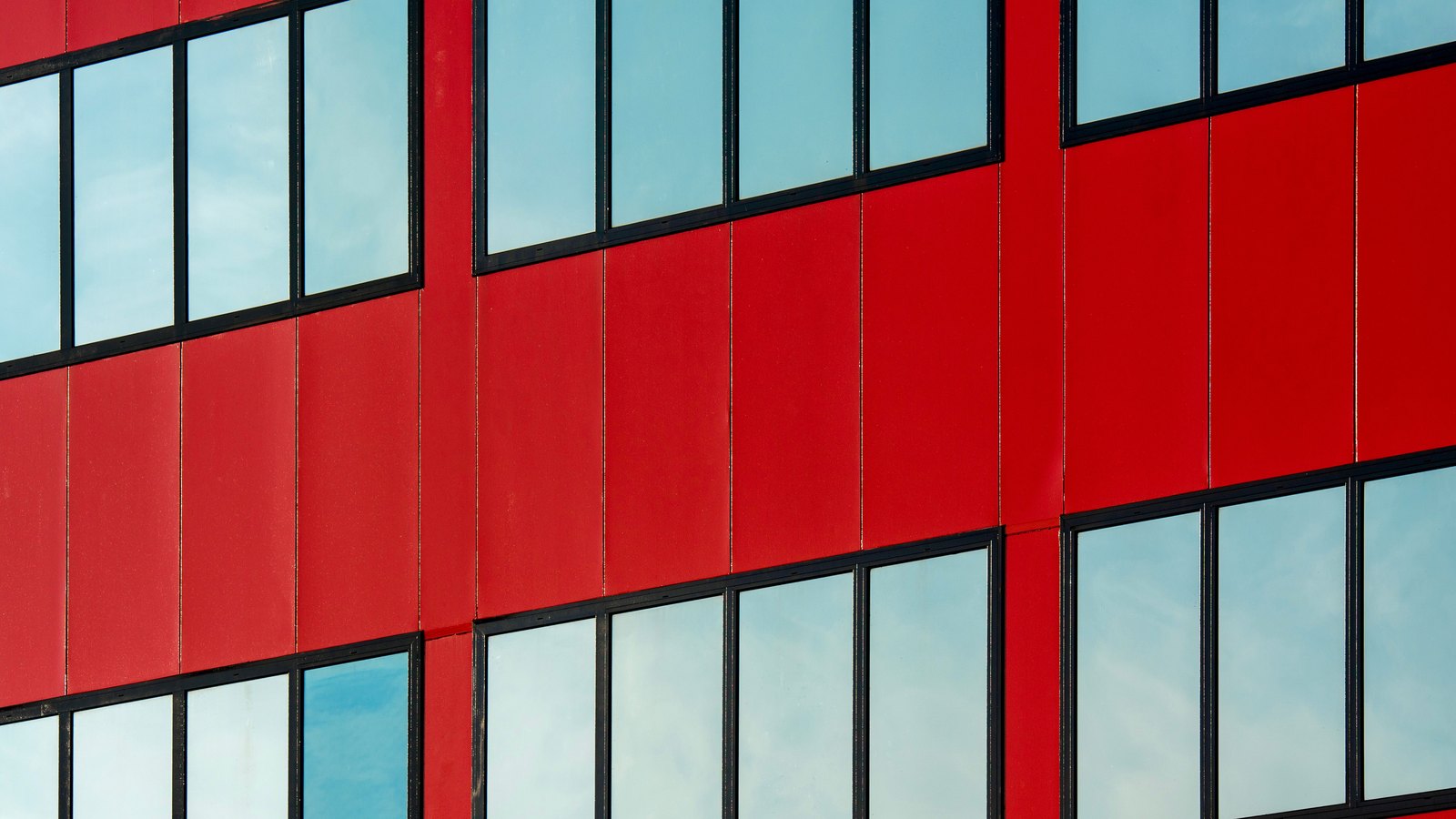 A close crop of the side of a modern building: flat scarlet cladding panels set in a grid and broken by rows of black framed windows that reflect pale sky and cloud. No lettering, sign, logo or person is in view.